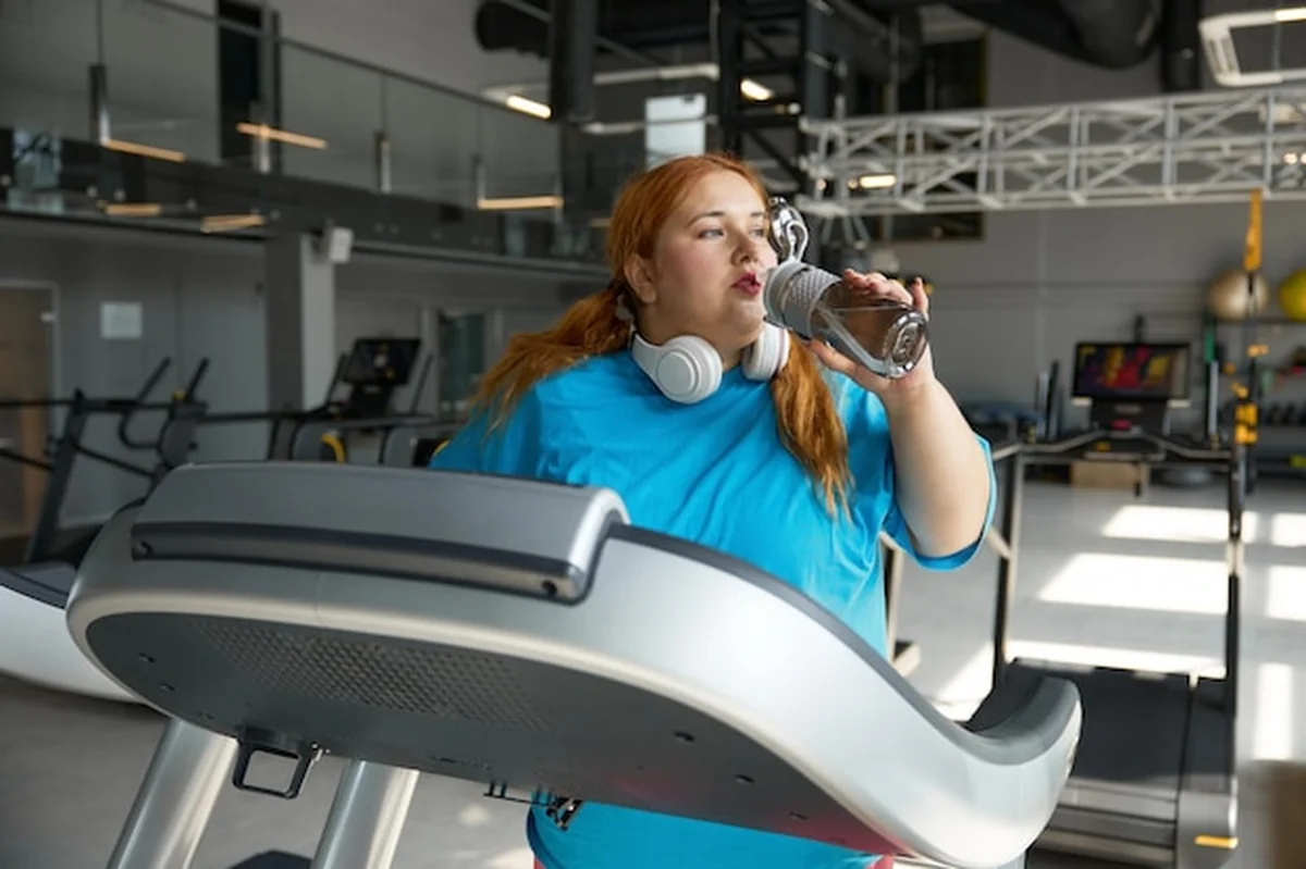 Wyczerpana sportowa kobieta z nadwagą pijąca wodę podczas treningu na bieżni w siłowni